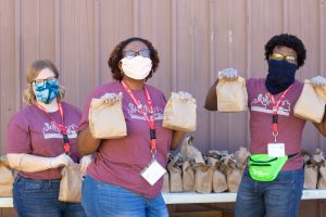 Three AmeriCorps members wearing cloth facemasks hold up brown bags during a meal distribution.